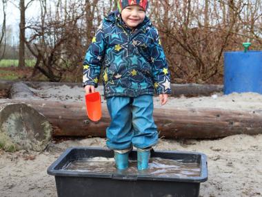 Dreng står i kæmpe kasse med vand og sand. Han nyder det, da han står med gummistøvler på.